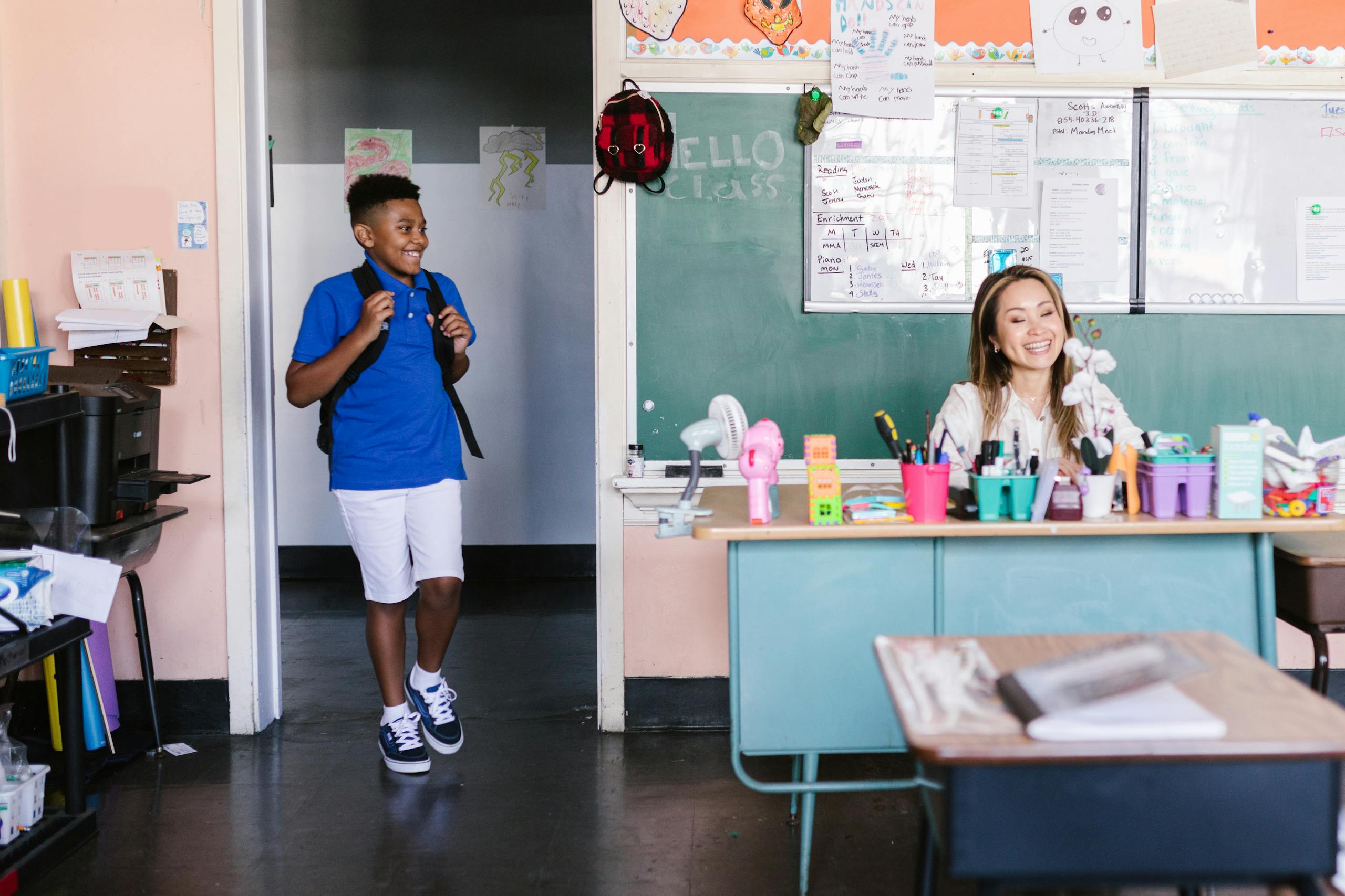 A cheerful student enters the classroom where a teacher is seated at a desk, ready for class.