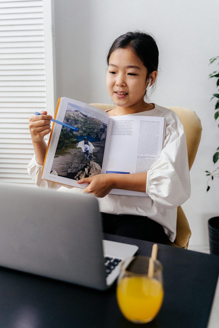 Asian girl showing an open book during a home virtual learning session with a laptop.